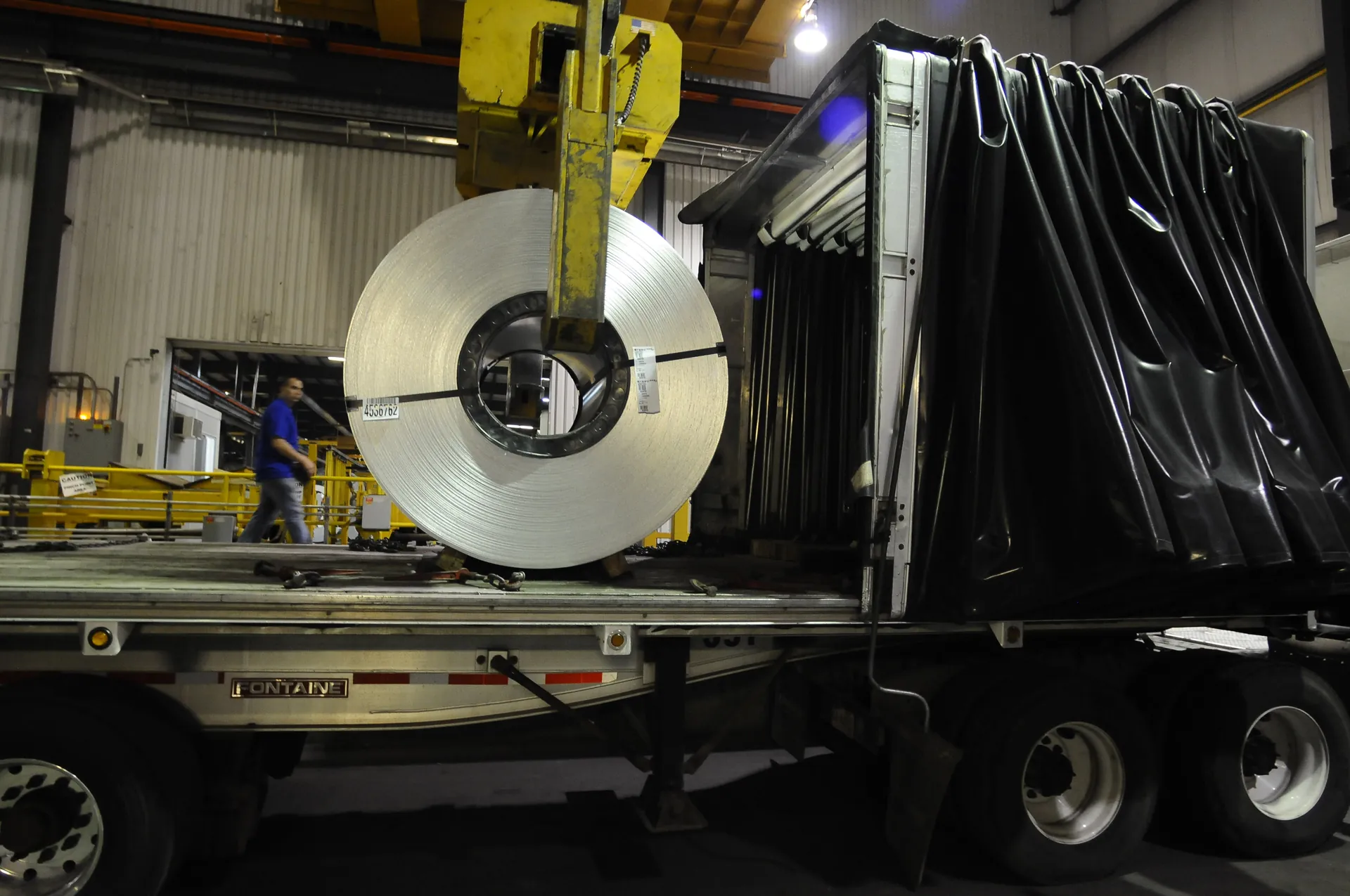 Crane C-hook lowering a steel coil onto a flatbed truck inside a service centre