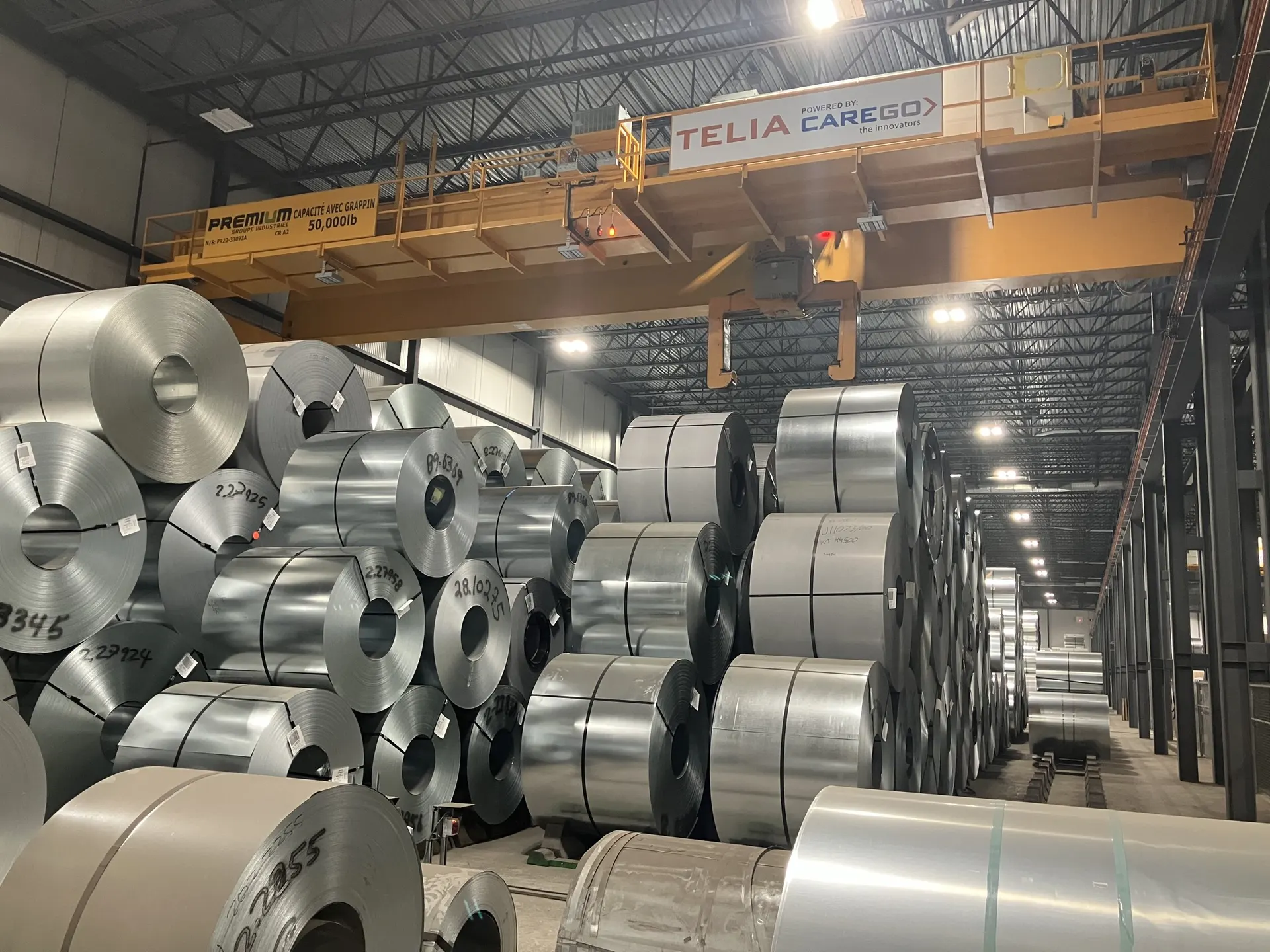 Overhead crane lifting a steel coil above a row of coils inside a mill